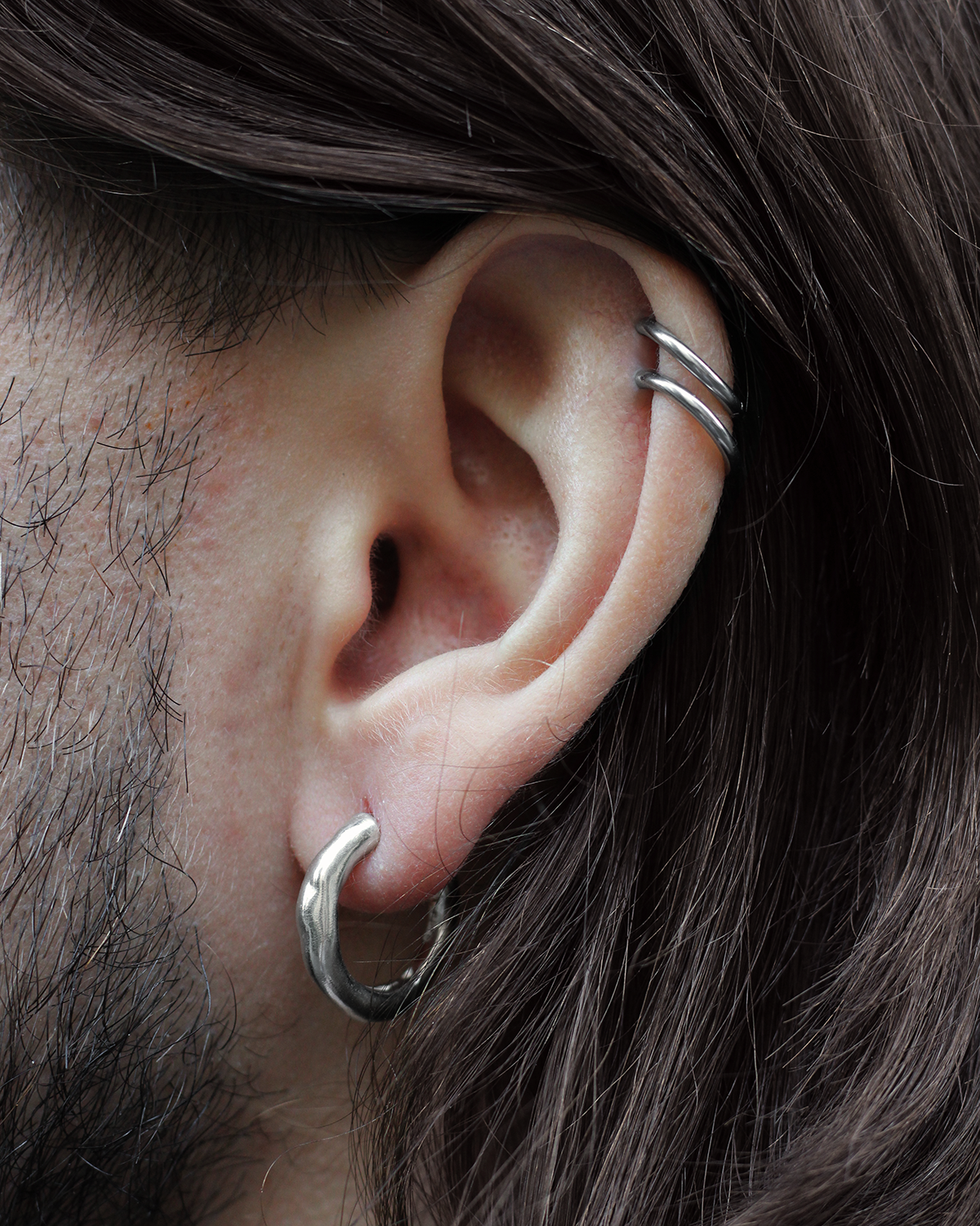 Close-up of a person's ear wearing a silver hoop earring in the lobe and two thin silver cartilage piercings, with dark brown hair and facial hair visible.