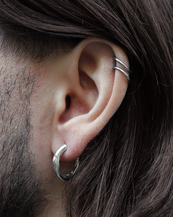 Close-up of a person's ear wearing a silver hoop earring in the lobe and two thin silver cartilage piercings, with dark brown hair and facial hair visible.